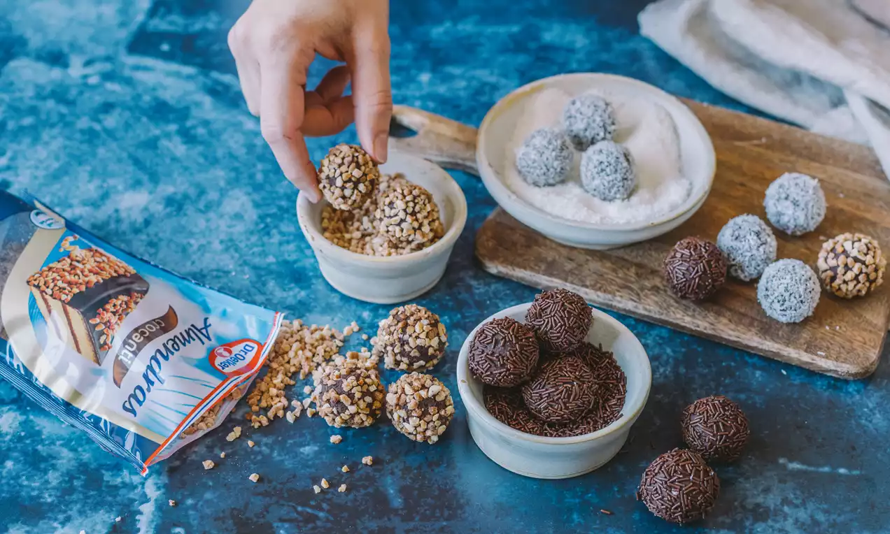 Trufas de chocolate redondas cubiertas con crocante de avellana, fideos de chocolate y coco rallado, dispuestas en cuencos y un plato sobre una mesa azul