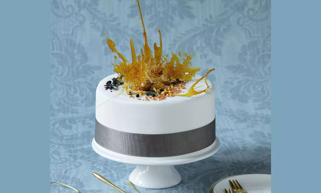 Kulový dort pokrytý hladkým bílým fondánem, s šedou stuhou, dekorovaný karamelovými střapci, zlatými perlami a černými drobky na podnosu