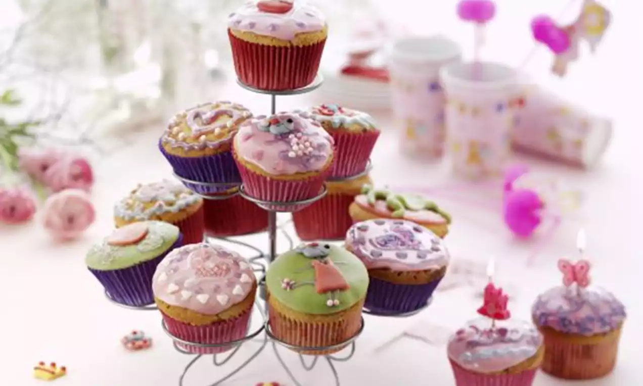 Cupcakes med glasur i pastellfarger og dekorative motiver på et metallstativ, omgitt av rosa pynt og lyse plastkopper i bakgrunnen