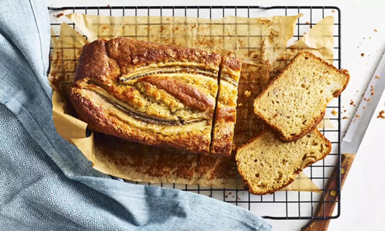 Golden-brown banana bread loaf with a glossy surface and baked banana slices, set on parchment paper atop a cooling rack with two slices cut