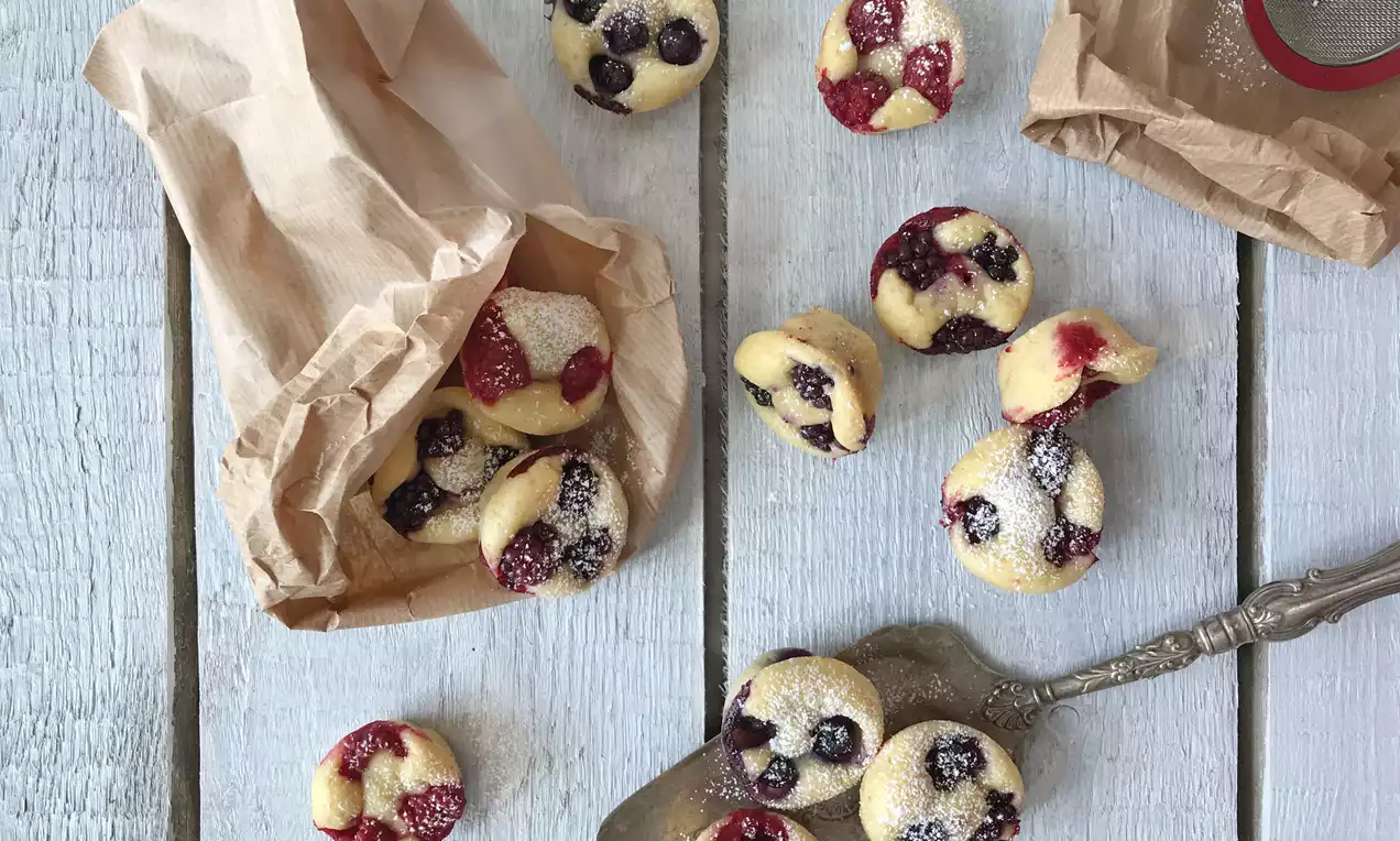 Pienet kullanvaaleat pannukakkupalat mustikalla ja vadelmalla, jauhoiselta paperipussilta lautaselle kaadettuina ja tomusokerilla siroteltuina