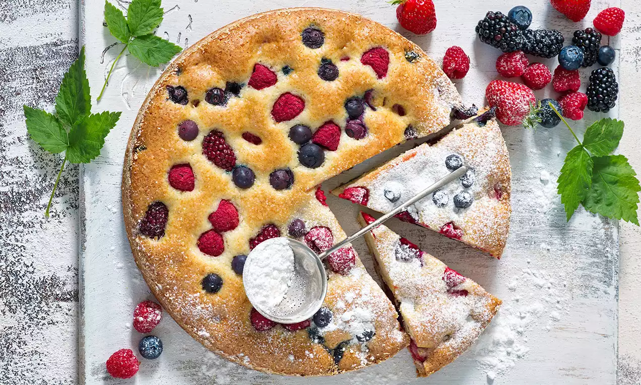 Zlatavá kulatá bublanina s posypem moučkového cukru, zdobená červenými a modrými lesními plody, servírovaná na světlém dřevěném podkladu