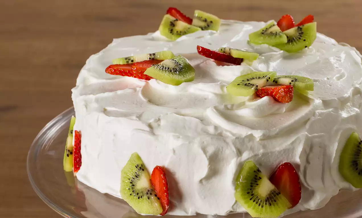 Torta soffice e bianca coperta da crema morbida, decorata con fette di kiwi verde brillante e fragole rosse su un piatto di vetro