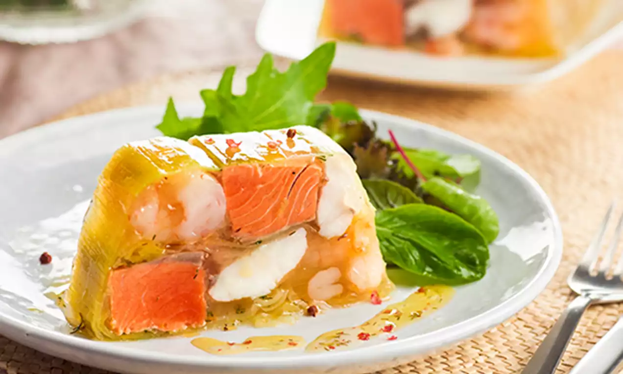 Terrina cubierta de gelatina dorada transparente con capas visibles de salmón naranja, gambas rosadas y huevo blanco, acompañada de hojas verdes