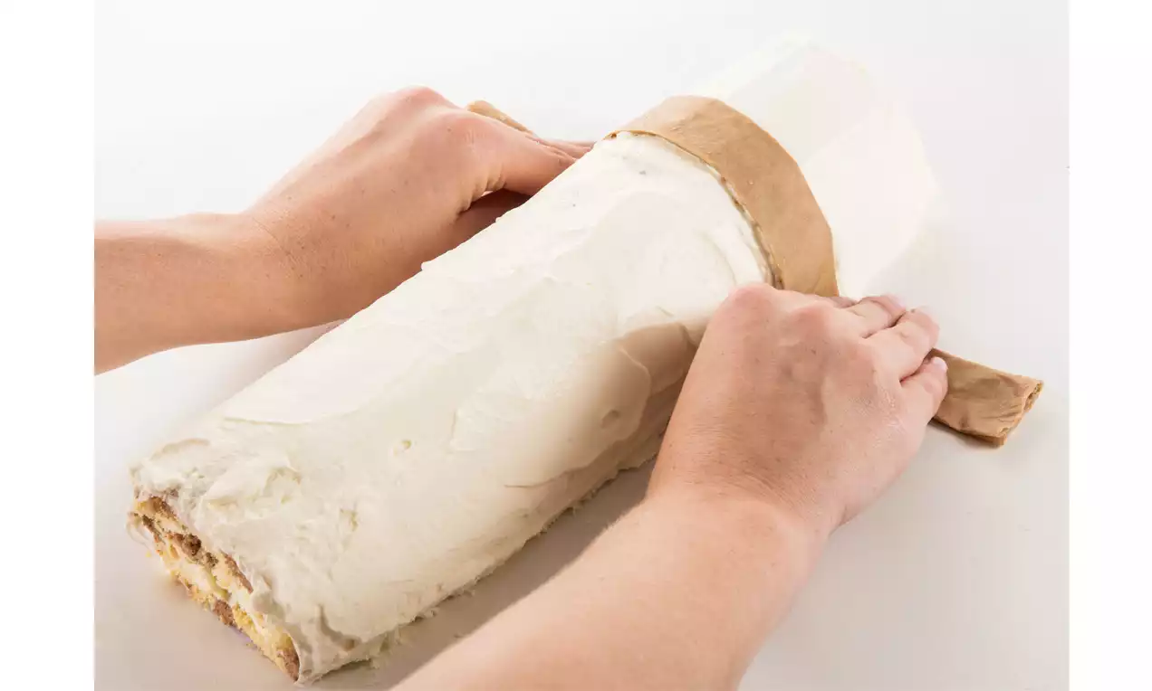 Helle Biskuitrolle mit glatter weißer Creme umhüllt, an den Enden leicht sichtbar gefüllt, von Händen mit braunem Papier eingerollt 
