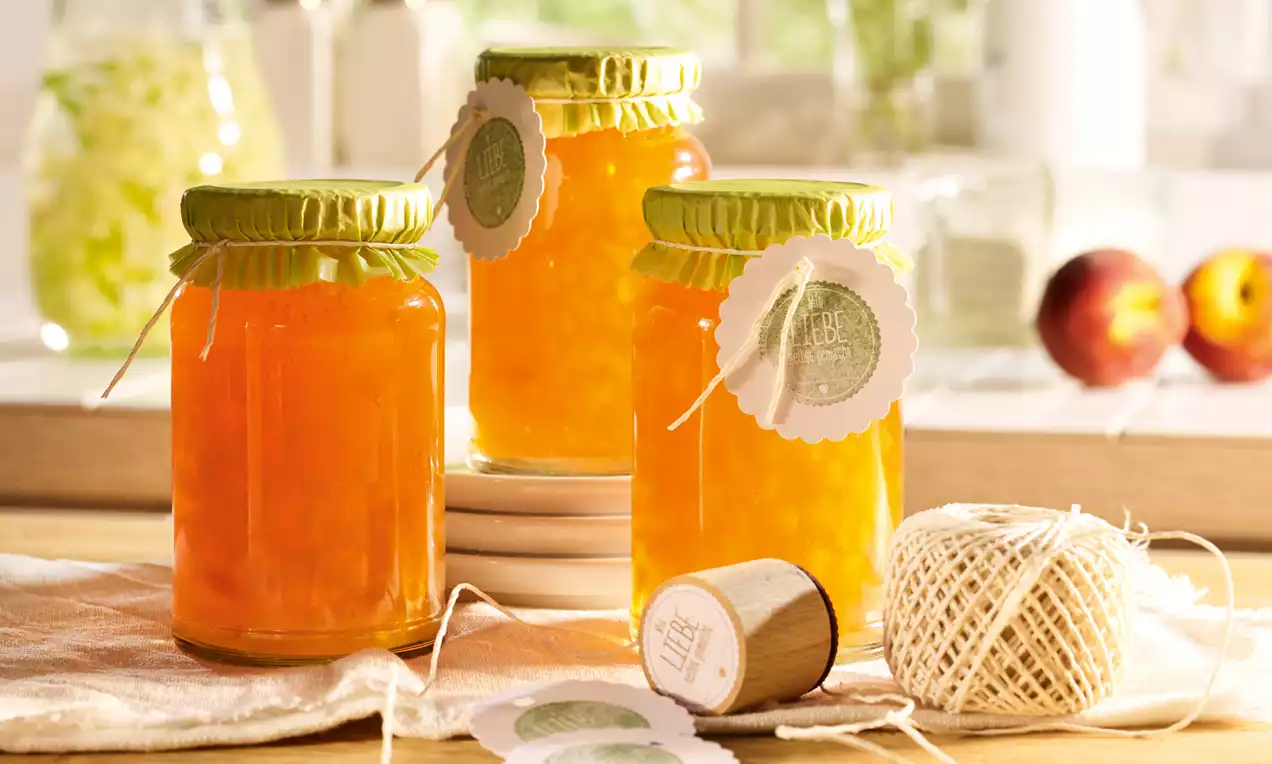 Gläser mit gold-orangem Holunderblüten-Apfel-Gelee und sichtbaren Pfirsichstückchen, mit grünen Deckeln und runden Anhängern verziert