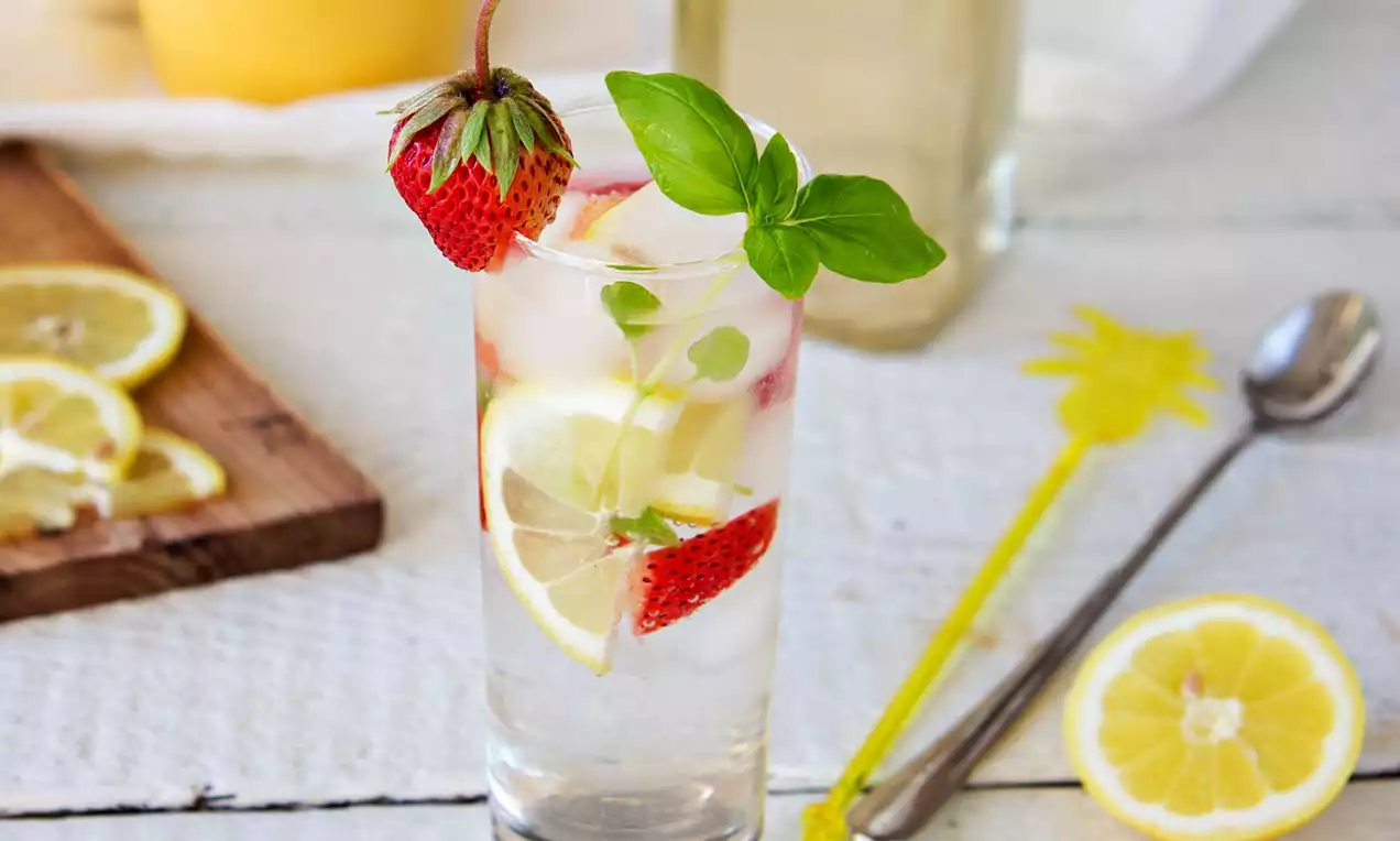 Průhledná sklenice plná světlého nápoje s plátky citronů, jahodami, kostkami ledu a čerstvými lístky bazalky na světlém dřevěném stole