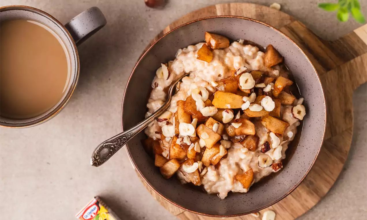 Šviesiai kreminės spalvos ryžių pudingas dubenėlyje, puoštas kepintais obuolių gabalėliais, lazdyno riešutais ir padėtas ant medinio padėklo