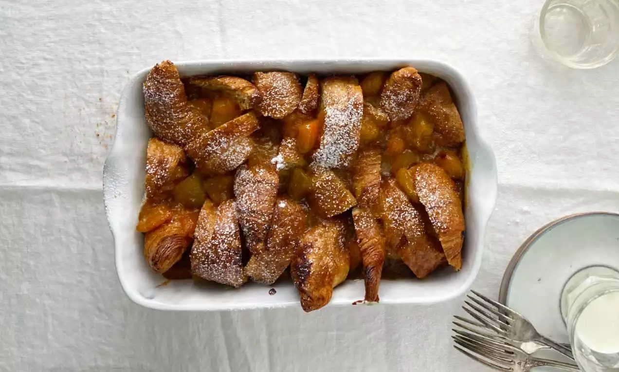 Croissants dorati e caramellati disposti in una teglia bianca, spolverati di zucchero a velo su uno sfondo di tovaglia candida