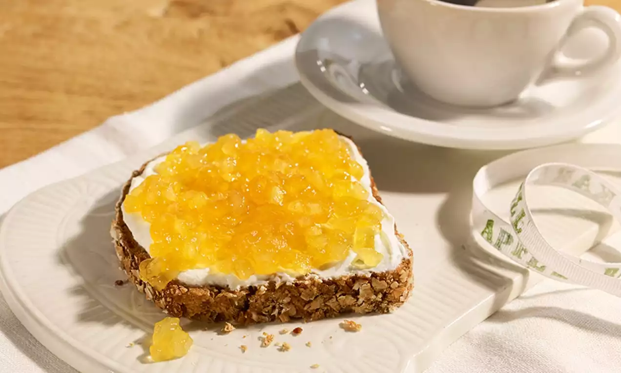 Dulceață de mere galben-aurie cu textură catifelată distribuită generos peste cremă albă pe o felie de pâine granuloasă, așezată pe o farfurie crem