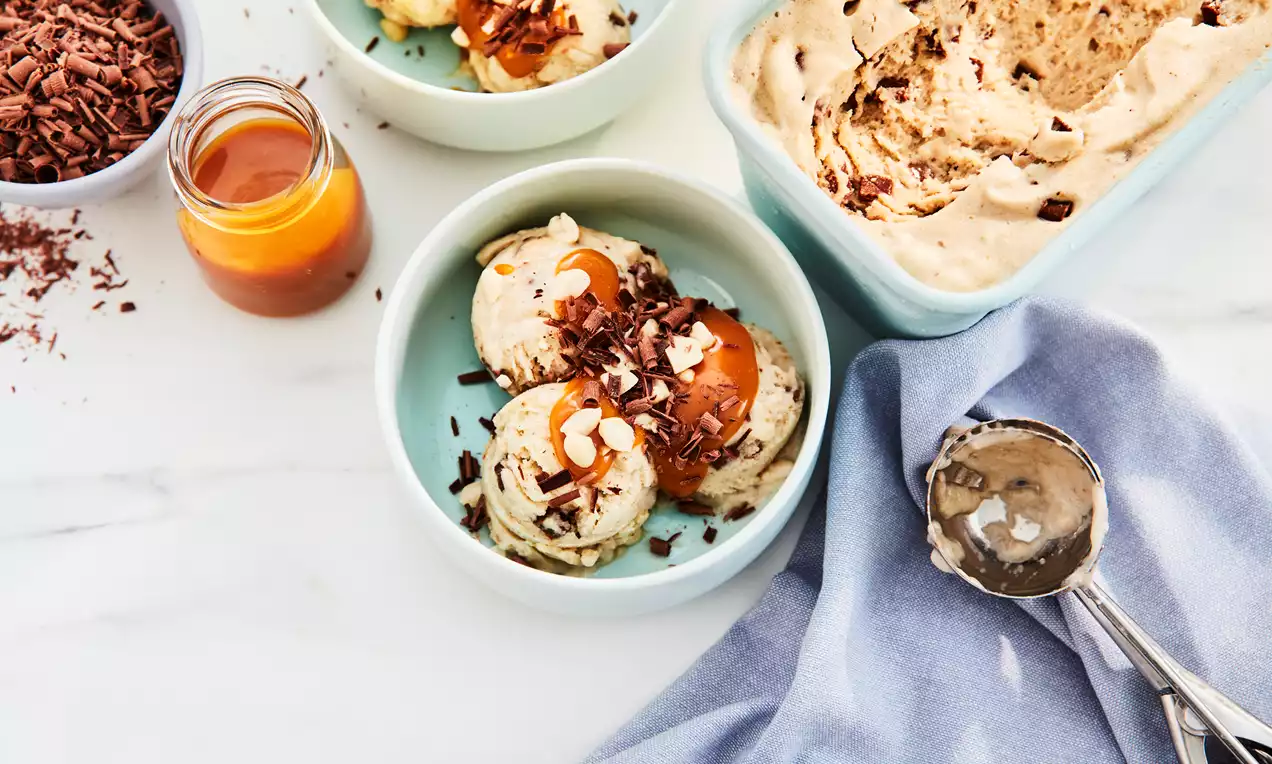 Banana ice cream pale and creamy, scooped into a mint-green bowl, topped with chocolate shavings and caramel sauce alongside a jar