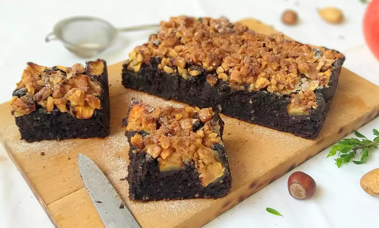 Karobový koláč tmavě hnědé barvy s kousky zlatavých jablek a křupavou ořechovou drobenkou na dřevěném prkénku doplněný lískovými oříšky