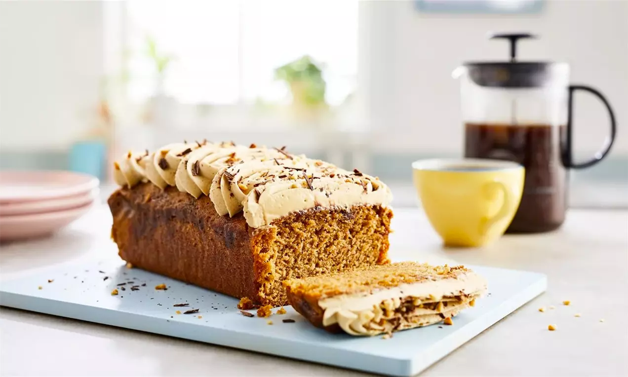 Правоъгълен кейк с тъмнокафяво тесто, покрит с рошав светложълт крем, декориран с шоколадови стърготини, разположен на светлосиня дъска