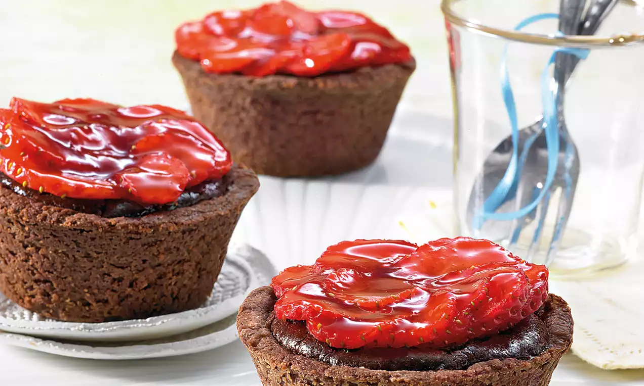 Čokoladne tortice tamnosmeđe boje s glatkim slojem sjajnog crvenog preljeva od jagoda, poslužene na bijelom tanjuru uz staklenu čašu i žličice