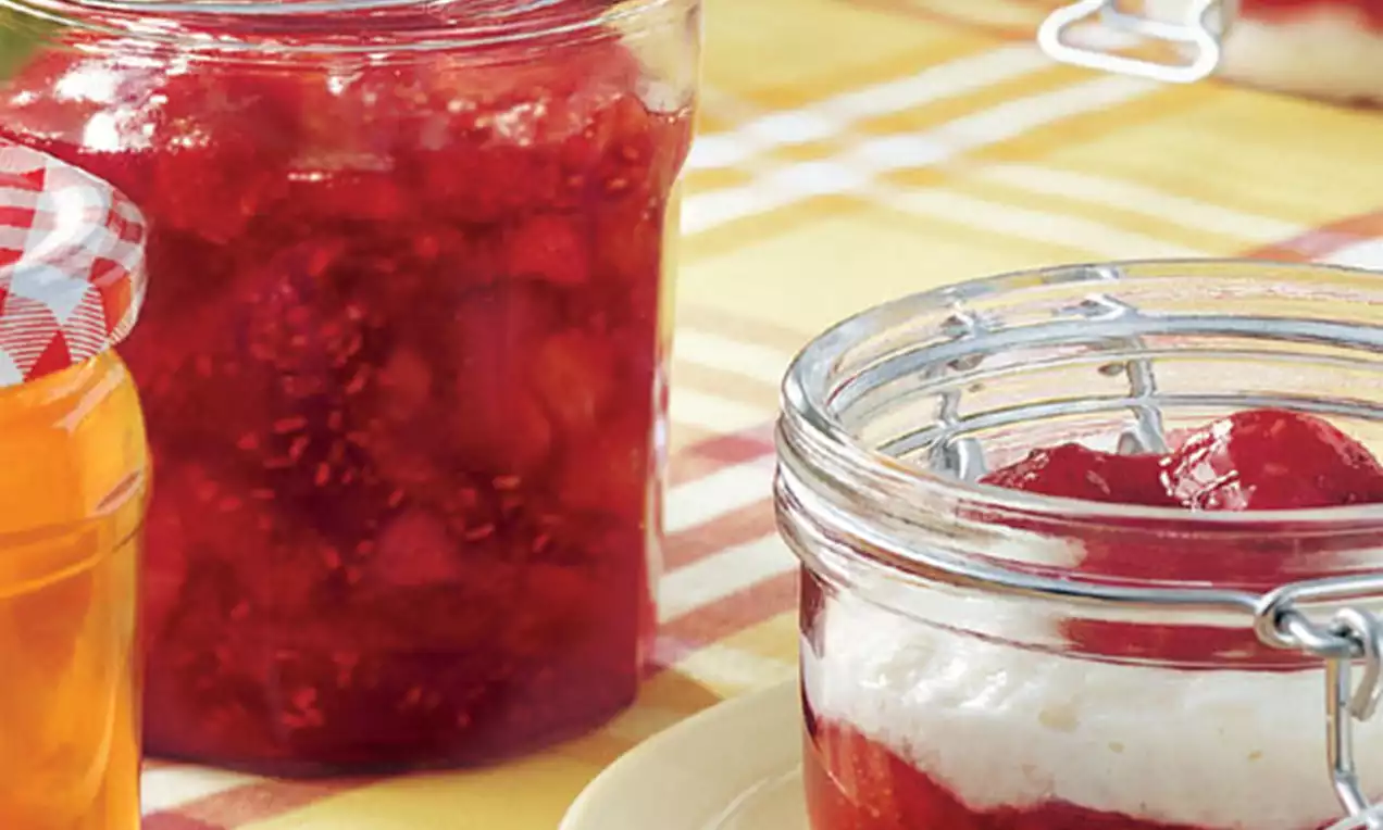 Glazen potten met glanzend rode aardbeienframbozenconfituur, grove textuur en fruitstukjes, gepresenteerd op een geel geruit tafelkleed