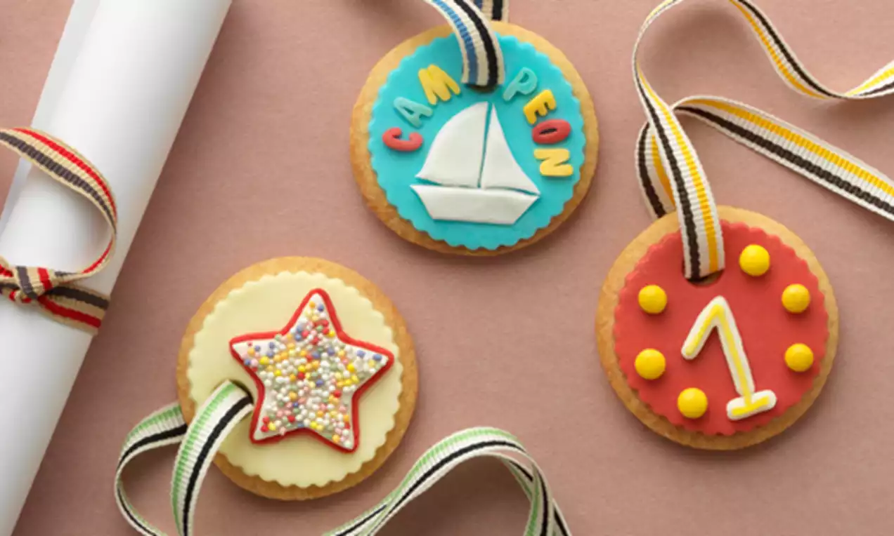 Medallas de galleta doradas decoradas con glaseado colorido, cintas rayadas y figuras de estrella, número y velero sobre fondo rosa