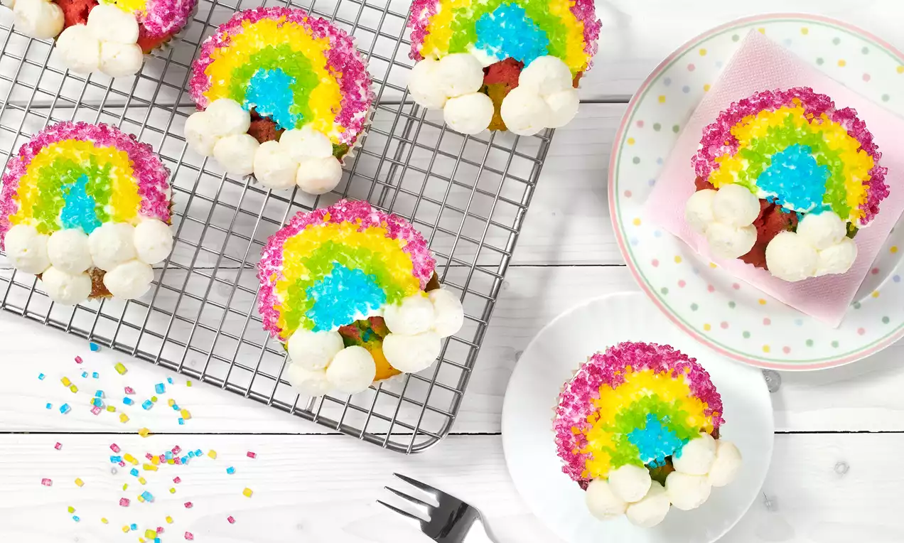 Bunte Regenbogen-Cupcakes mit farbigem Zuckerguss, weißen Sahnewolken und pinken Zuckerstreuseln auf einer Abkühlgitter und Tellern angerichtet