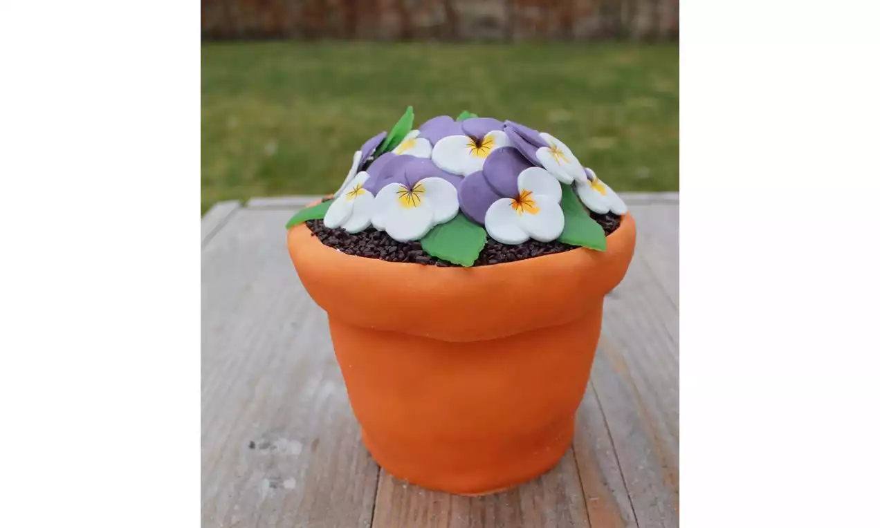 Kake formet som en terrakottapotte med brun sjokoladestrø på toppen, pyntet med lilla og hvite blomster i fondant med grønne blader