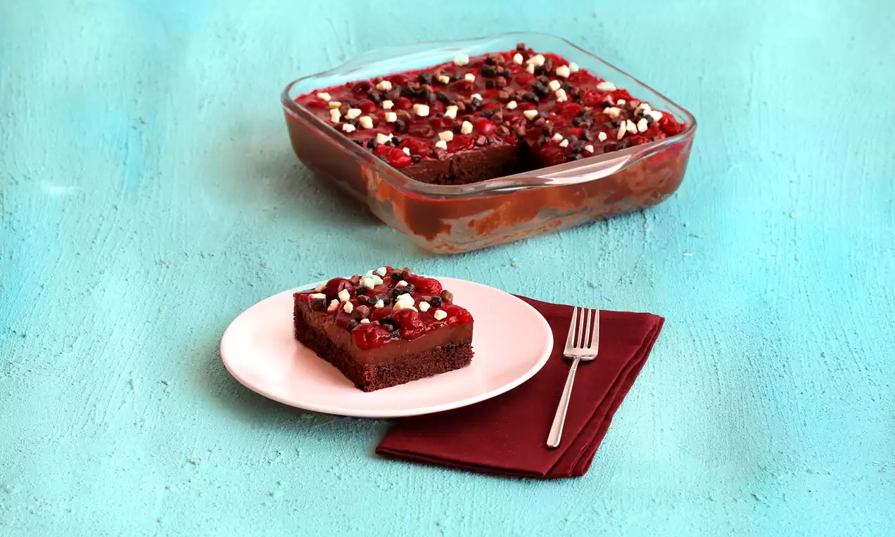 Cam bir kapta dilimlenmiş, üstü vişne taneleri ve beyaz çikolata parçalarıyla süslü kakaolu kek, beyaz tabakta ve bordo peçeteyle sunulmuş