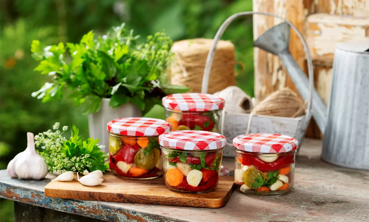 Murături colorate cu roșii verzi, ardei roșii și rondele de morcovi în borcane cu capac în carouri roșu-albe, așezate pe o masă rustică