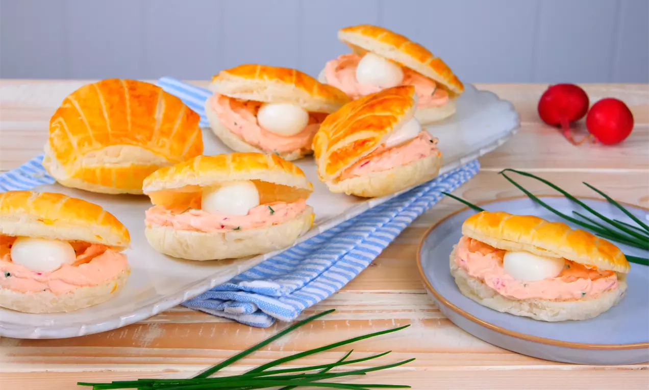 Goldgelbe, halbierte Blätterteigmuscheln mit cremiger, lachsfarbener Füllung und eingelegten Mini-Mozzarellakugeln auf einem Tablett angerichtet