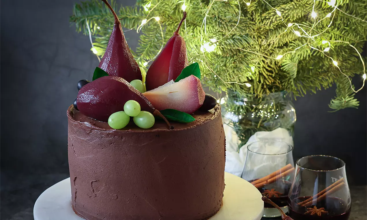 Шоколадова кръгла торта с гладка повърхност, увенчана с червени поширани круши, зелено грозде и листа върху бяла чиния