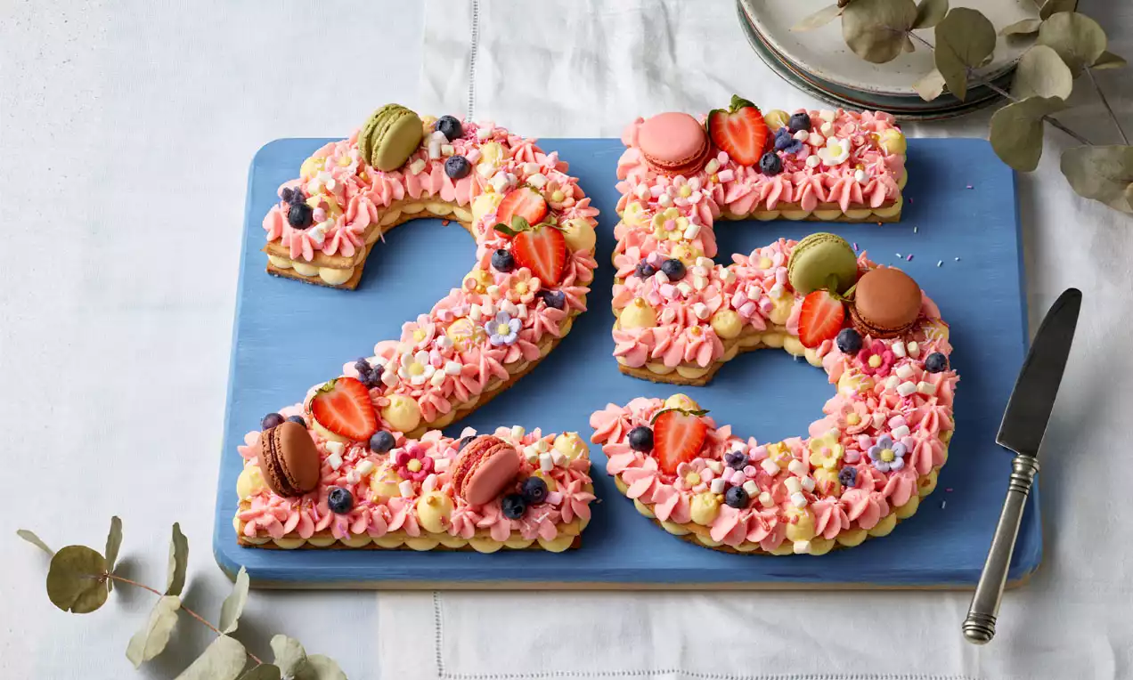 Zahlentorte in Form einer 25 aus hellem Mürbeteig mit rosafarbenen Cremetupfen, frischen Früchten, bunten Macarons und Perlen auf blauer Platte