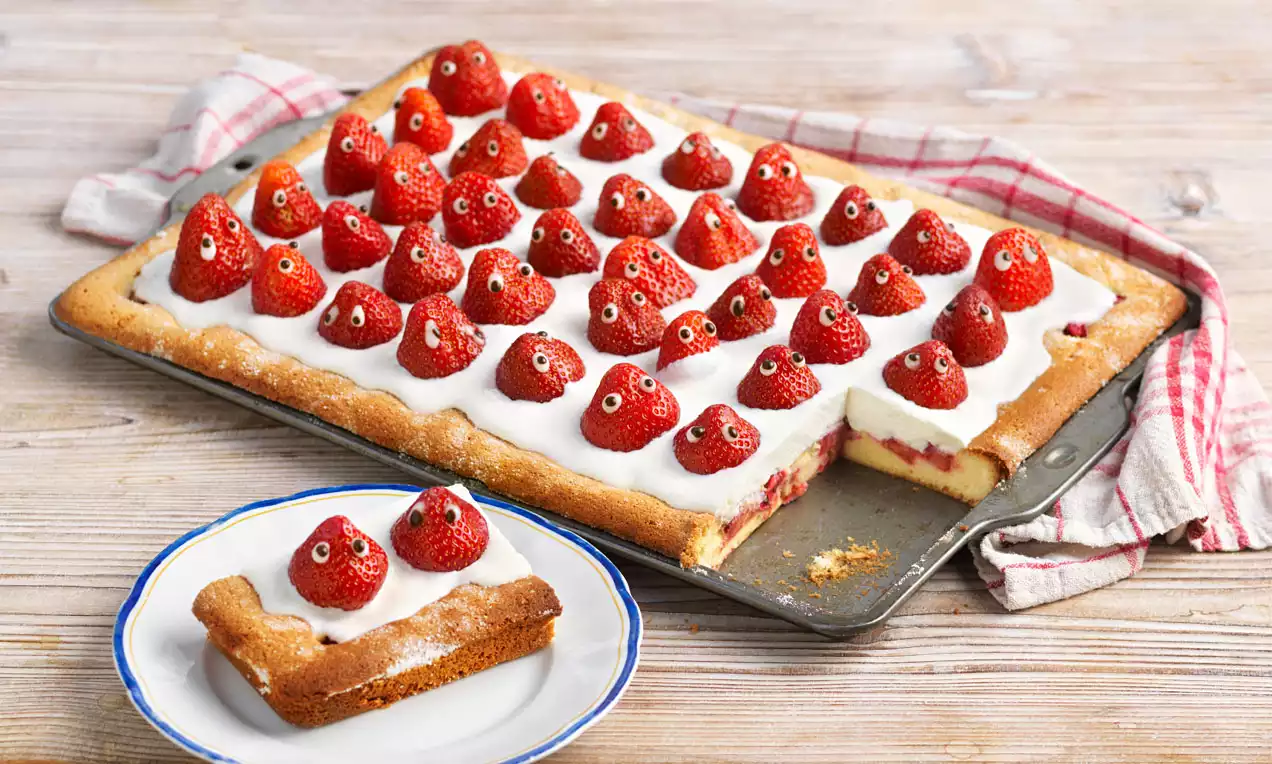 Rechteckiger Blechkuchen mit hellbraunem Boden, weißer Creme und ganzen Erdbeeren mit Zuckeraugen, ein Stück auf einem kleinen Teller