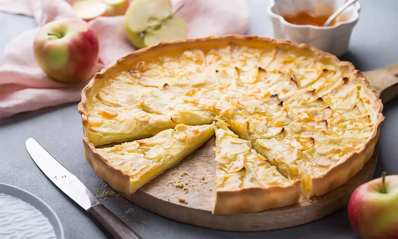 Goldgelbe Apfeltarte mit dünnen Apfelscheiben auf knusprigem Mürbeteig, angeschnitten auf einem Holzbrett, umgeben von frischen Äpfeln und Apfelmus