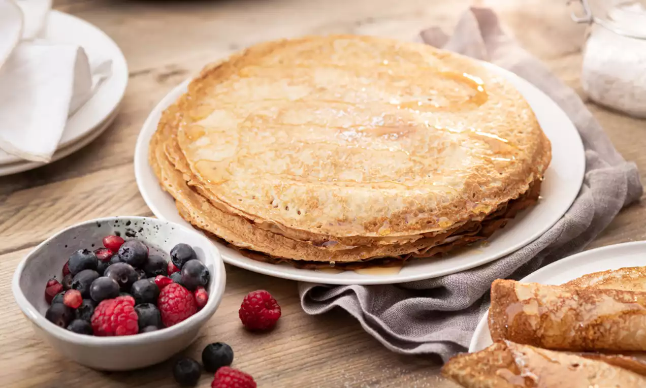 Crêpes dorées et moelleuses empilées sur une assiette blanche, accompagnées d’un bol de fruits rouges et noirs sur une table en bois