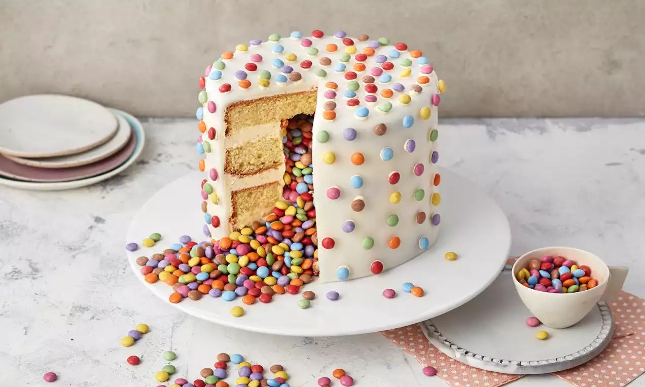 Hoher, weißer Überraschungskuchen mit bunten Schokolinsen dekoriert, gefüllt mit farbigen Linsen, auf weißer Tortenplatte angerichtet