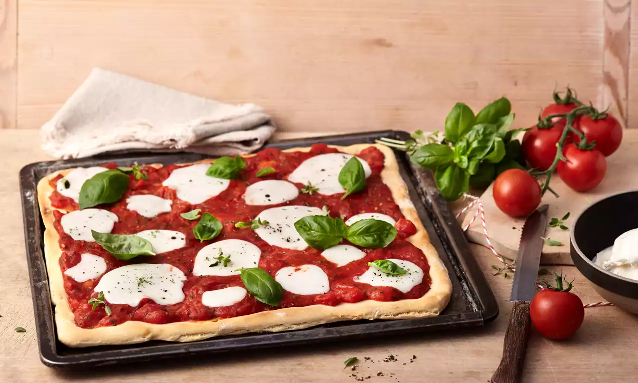 Pizzateig auf bemehltem Holzbrett, dünn ausgerollt und belegt mit roter Tomatensauce, Mozzarella und grünen Basilikumblättern