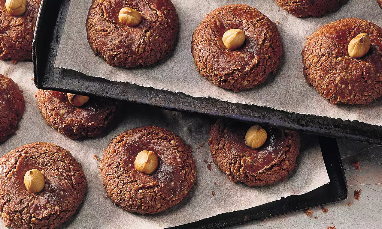 Auf einem Backblech braune, runde Kekse mit leicht rissiger Oberfläche, in der Mitte mit einer ganzen Haselnuss verziert, auf Backpapier