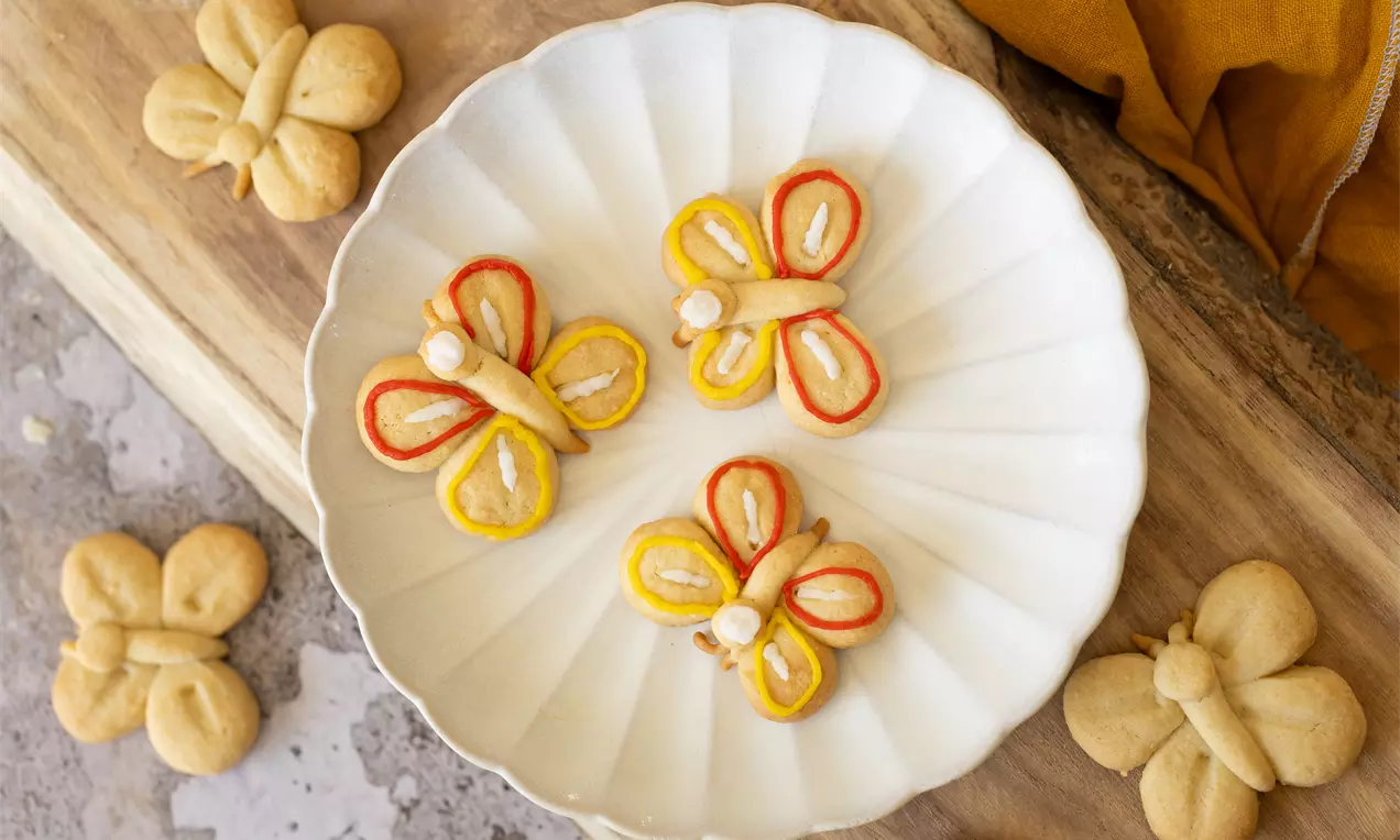 Biscuits en forme de papillons ornés de contours glacés orange et jaunes, disposés sur une assiette blanche nervurée en céramique