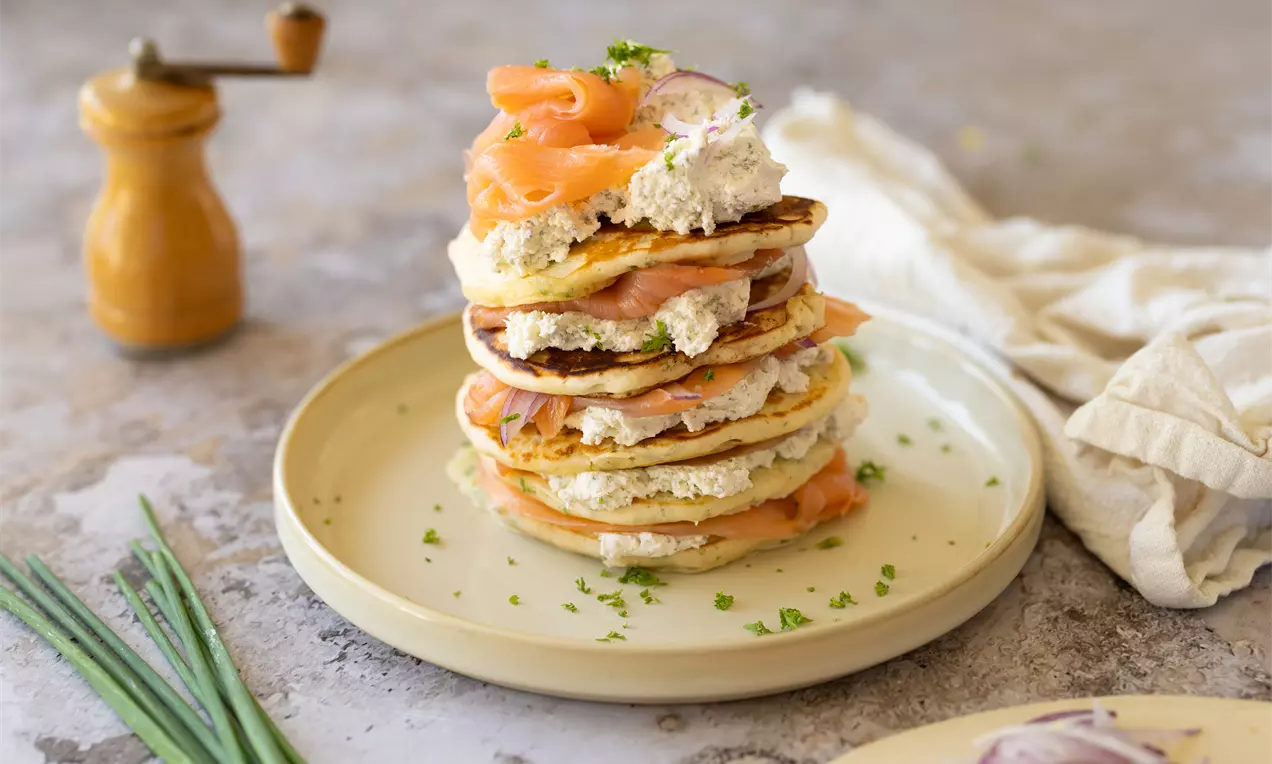 Een toren van goudbruine pancakes met zalmroosjes, romige witte topping en groene kruiden op een rond beige bord