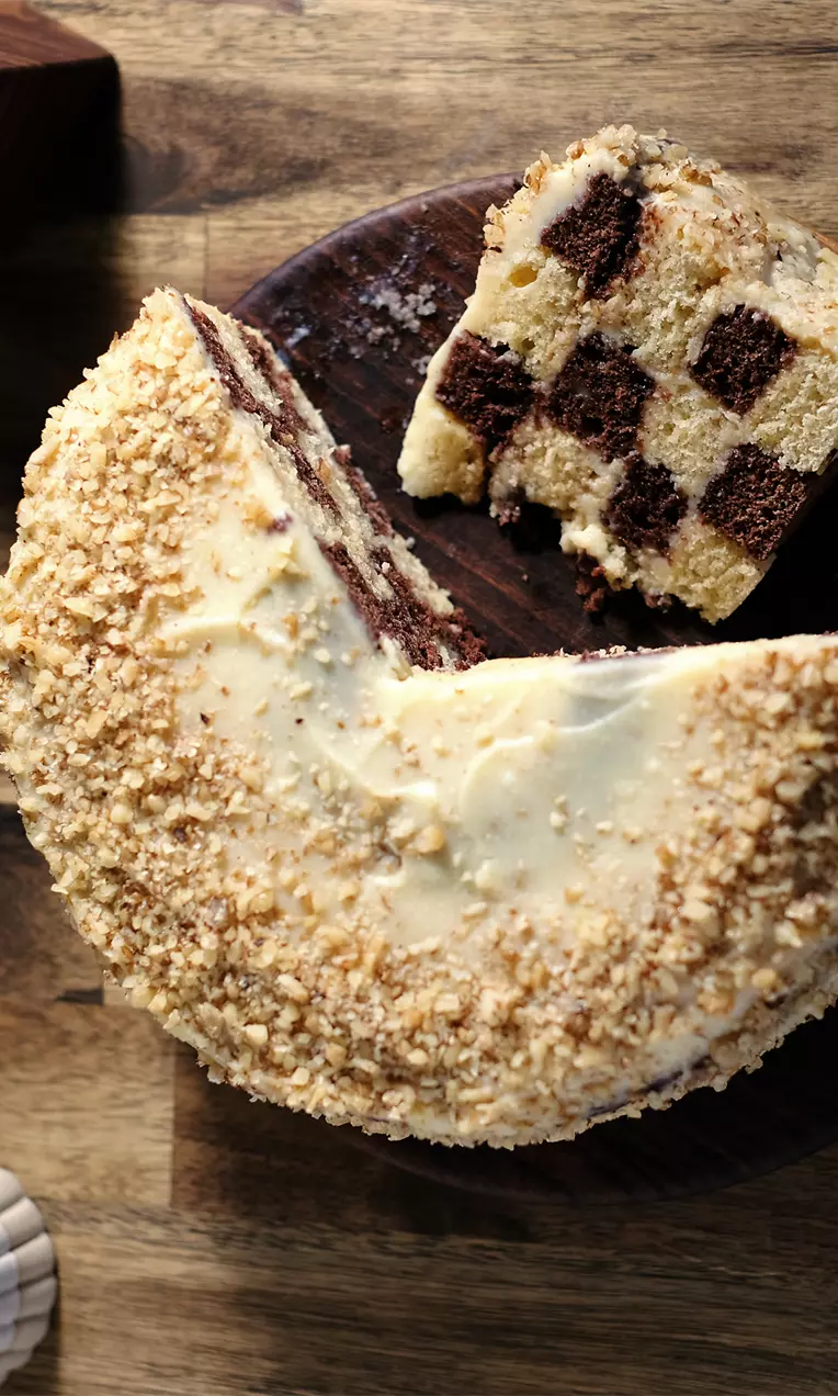 Кръгла торта с външна коричка от натрошени ядки, вътрешно редуващи се квадратчета светъл и тъмен блат, поднесена върху дървена плоча