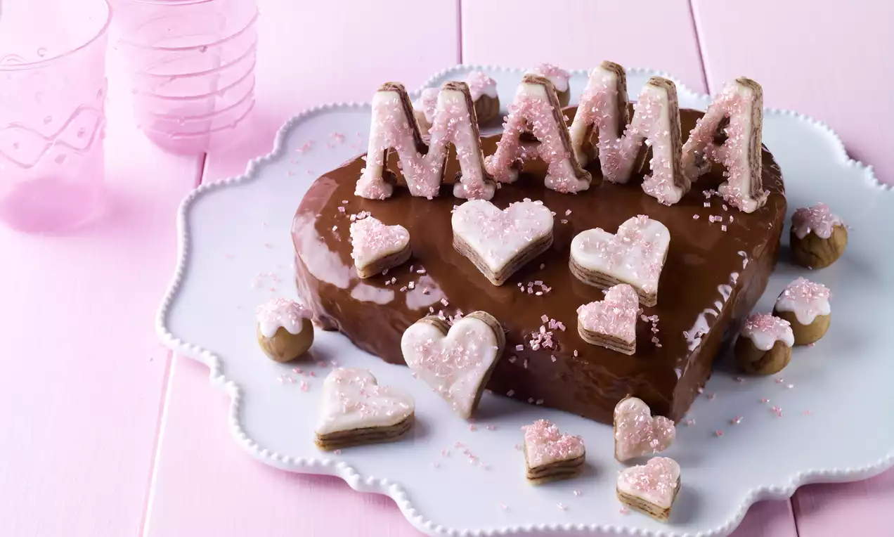 Ein herzförmiger Kuchen mit glänzender Schokoladenglasur, bestreut mit rosa Zucker, Marzipanbuchstaben und Herzdekoration auf weißer Tortenplatte