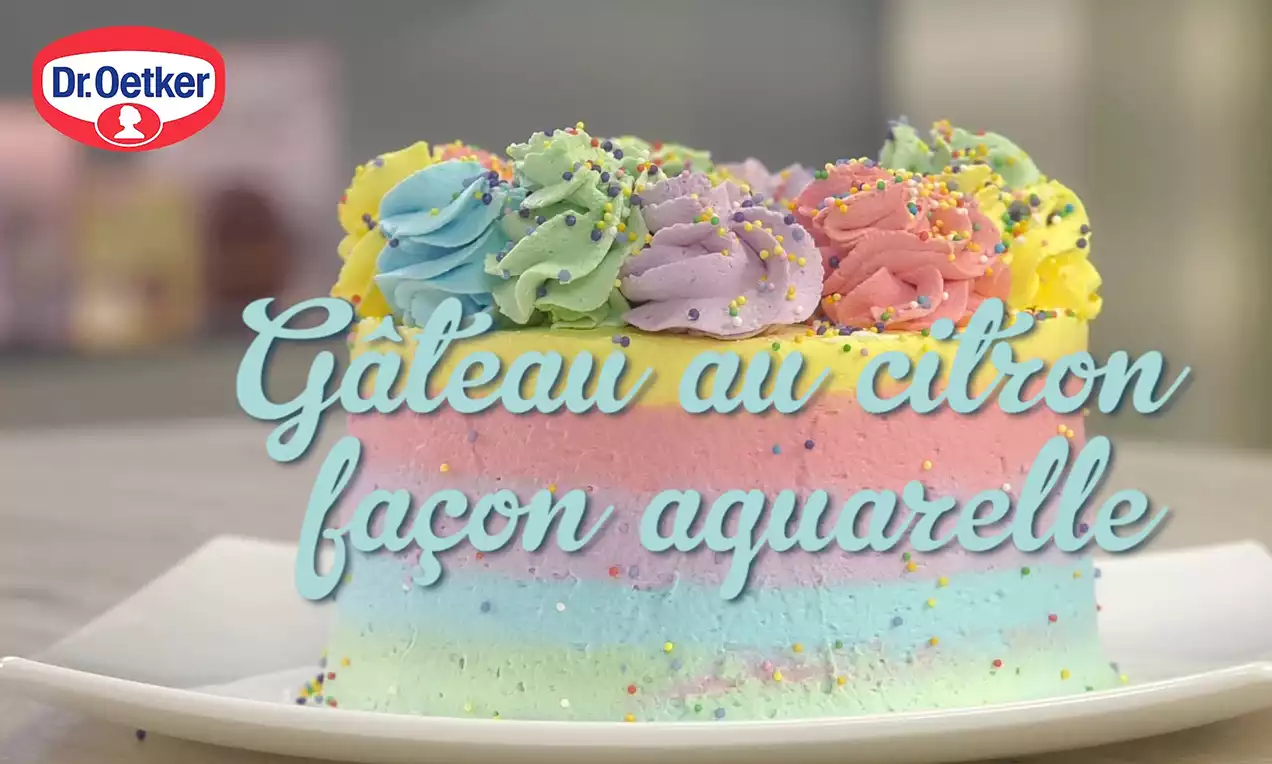 Gâteau aux couleurs pastel dégradées bleu, rose et jaune, couronné de rosaces multicolores et parsemé de petits éclats colorés sur une assiette blanche