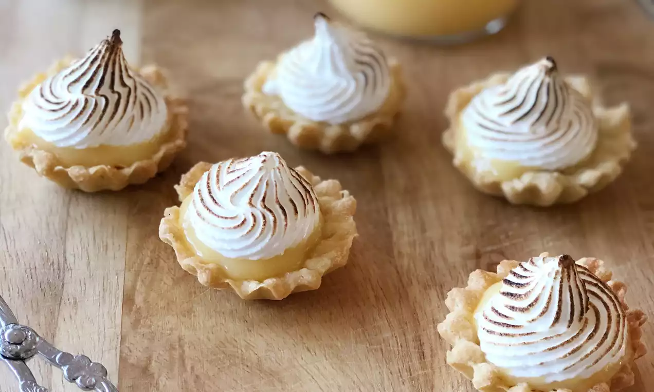 Ljusbruna minimarängpajer med gyllene kanter och toppar av vitbrunbränd maräng på träbord bredvid burk med gul citronkräm och tång