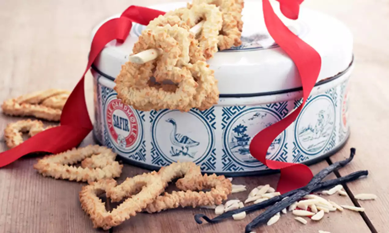 Sprø vaniljehjerter i lys gyllen farge, noen henger på en blå og hvit kakeboks med rødt bånd, omgitt av vaniljestenger og mandelskiver