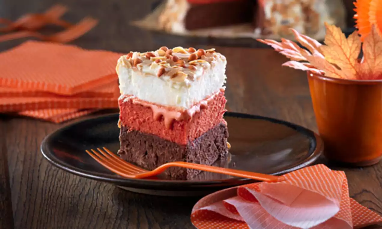 Porción triangular de tarta de tres capas, con base marrón de chocolate, centro rojo y cima blanca salpicada de frutos secos, servida en un plato oscuro junto a un tenedor naranja y servilletas a juego