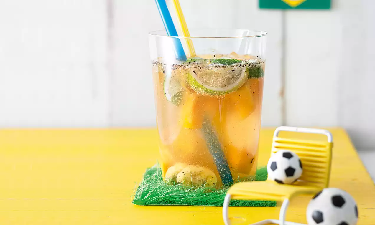 Ein durchsichtiger Glasbecher mit goldgelber Flüssigkeit, Limettenscheiben, Eiswürfeln, blauen und gelben Trinkhalmen auf gelber Tischplatte