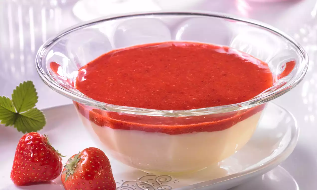 Glas mit zweilagigem Dessert aus weißem Sahnepudding und glänzendem rotem Erdbeerpüree, daneben frische Erdbeeren auf einem Teller