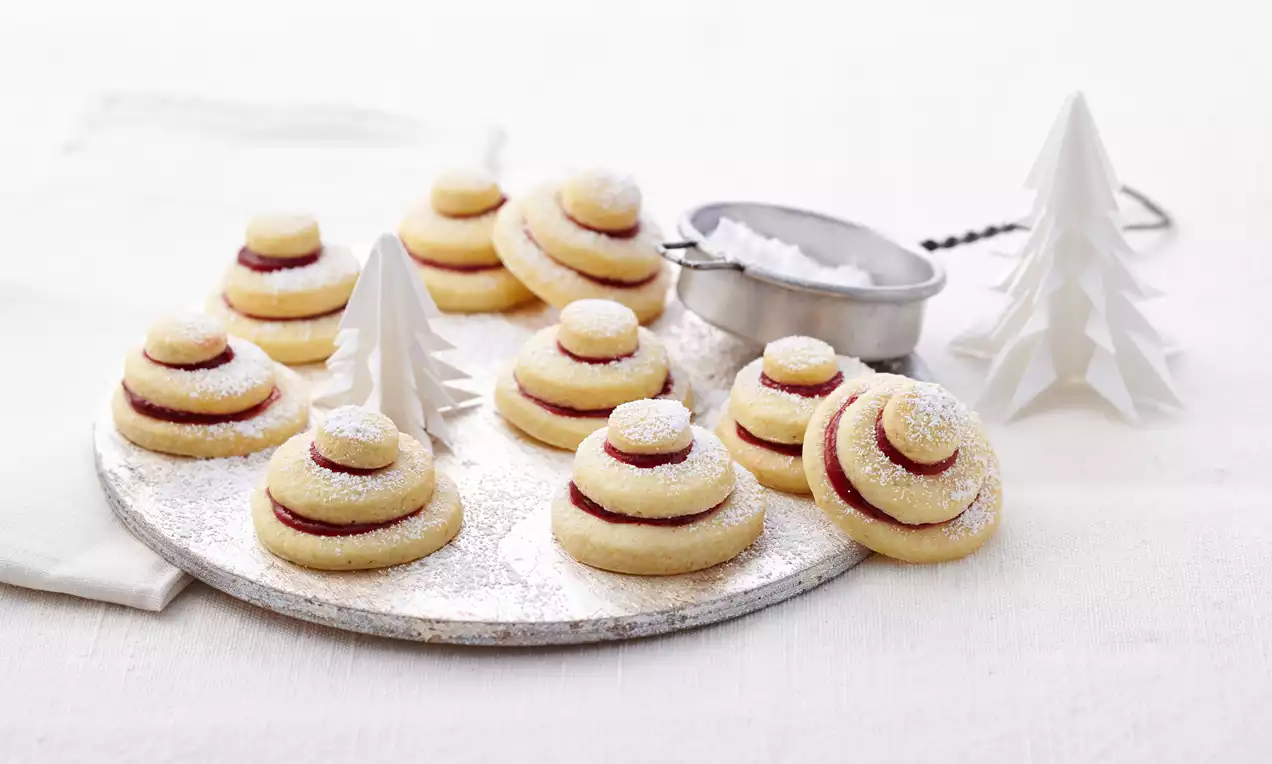 Terrassenplätzchen aus hellgoldenem Mürbeteig mit roter Füllung und Puderzucker bestäubt auf einem weißen Teller mit Papierdeko und Sieb
