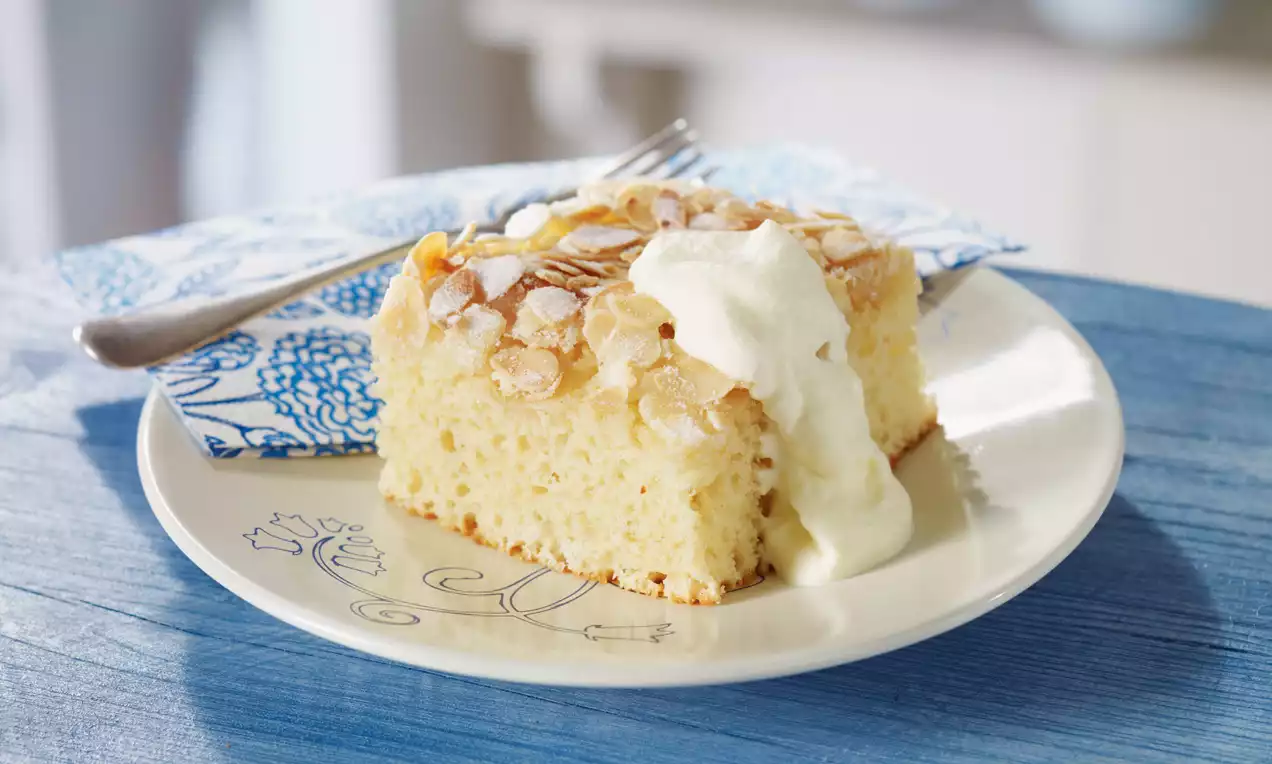 Carré de gâteau moelleux jaune doré garni d'amandes effilées et crème blanche, posé sur une assiette blanche décorée