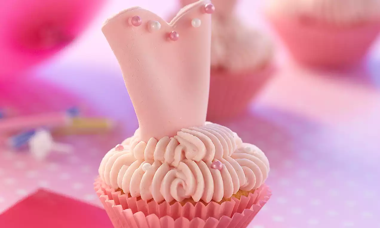 Cupcake con base circolare e crema rosa a spirali, decorato con un topper a forma di tutù rosa ornato da perline colorate
