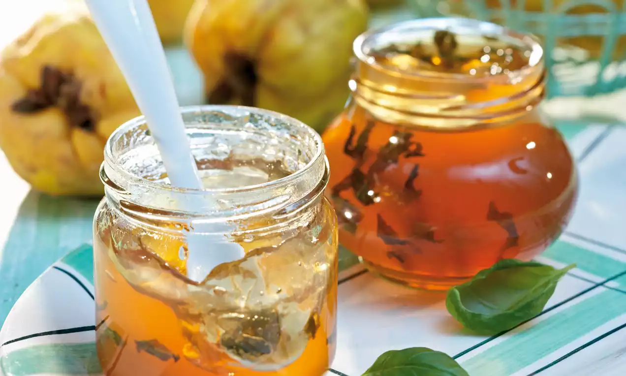 Goldbraunes Quittengelee mit schimmernder Oberfläche und Basilikumblättern in zwei Glasbehältern, umgeben von frischen Quitten und Basilikum