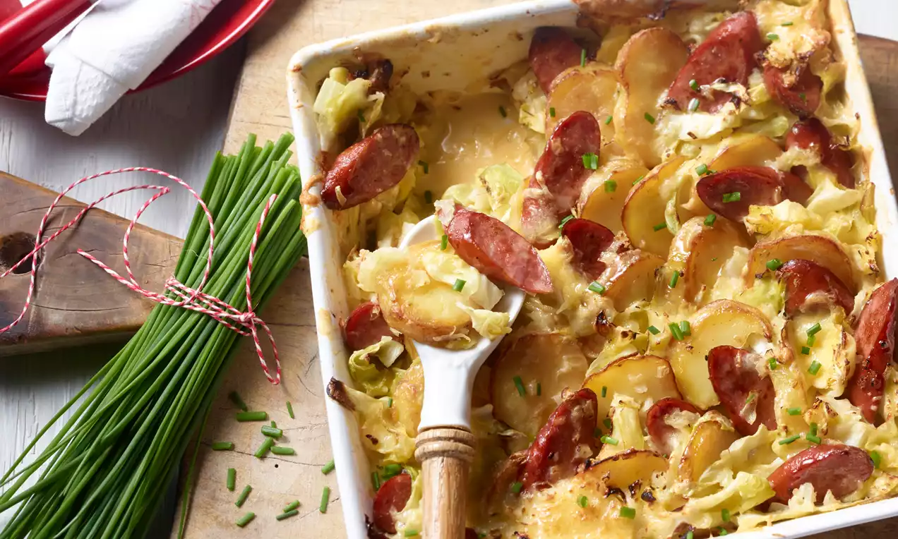 Goldbraune Bratkartoffelscheiben mit roten Wurststücken und grünen Schnittlauchringen in cremiger Sauce in einer weißen Auflaufform