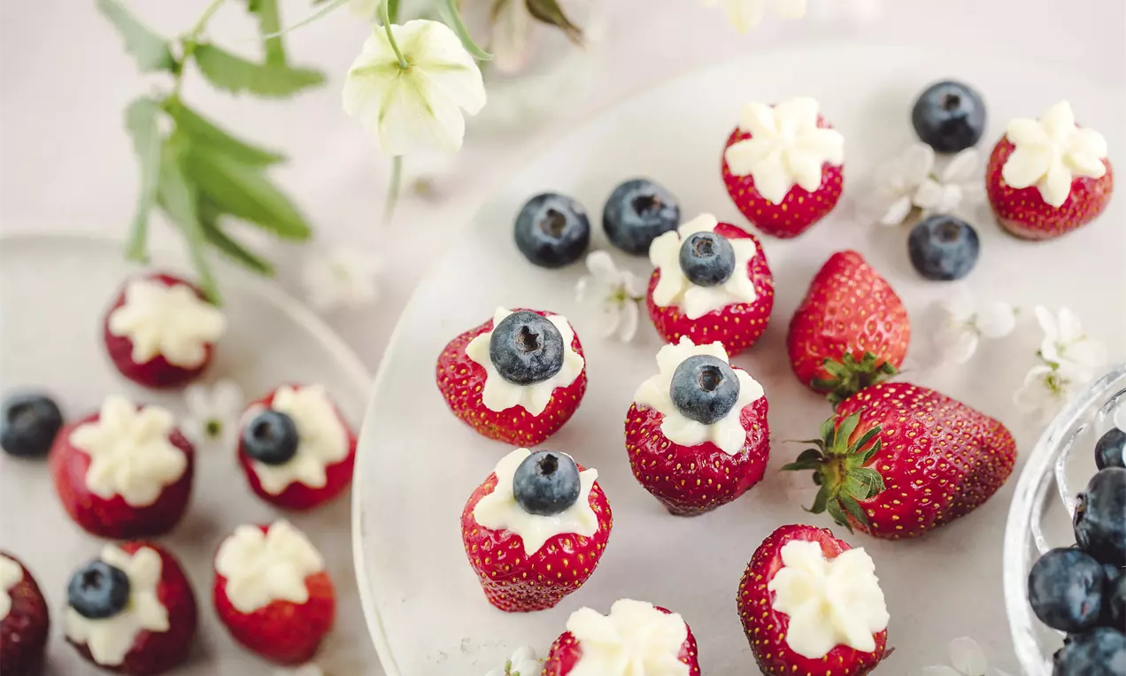 Svaigas, sarkanas zemeņu pusītes ar gludu krēmveida pildījumu un sulīgu melleņu akcentu, izkārtotas uz gaiša šķīvja blakus ziediem