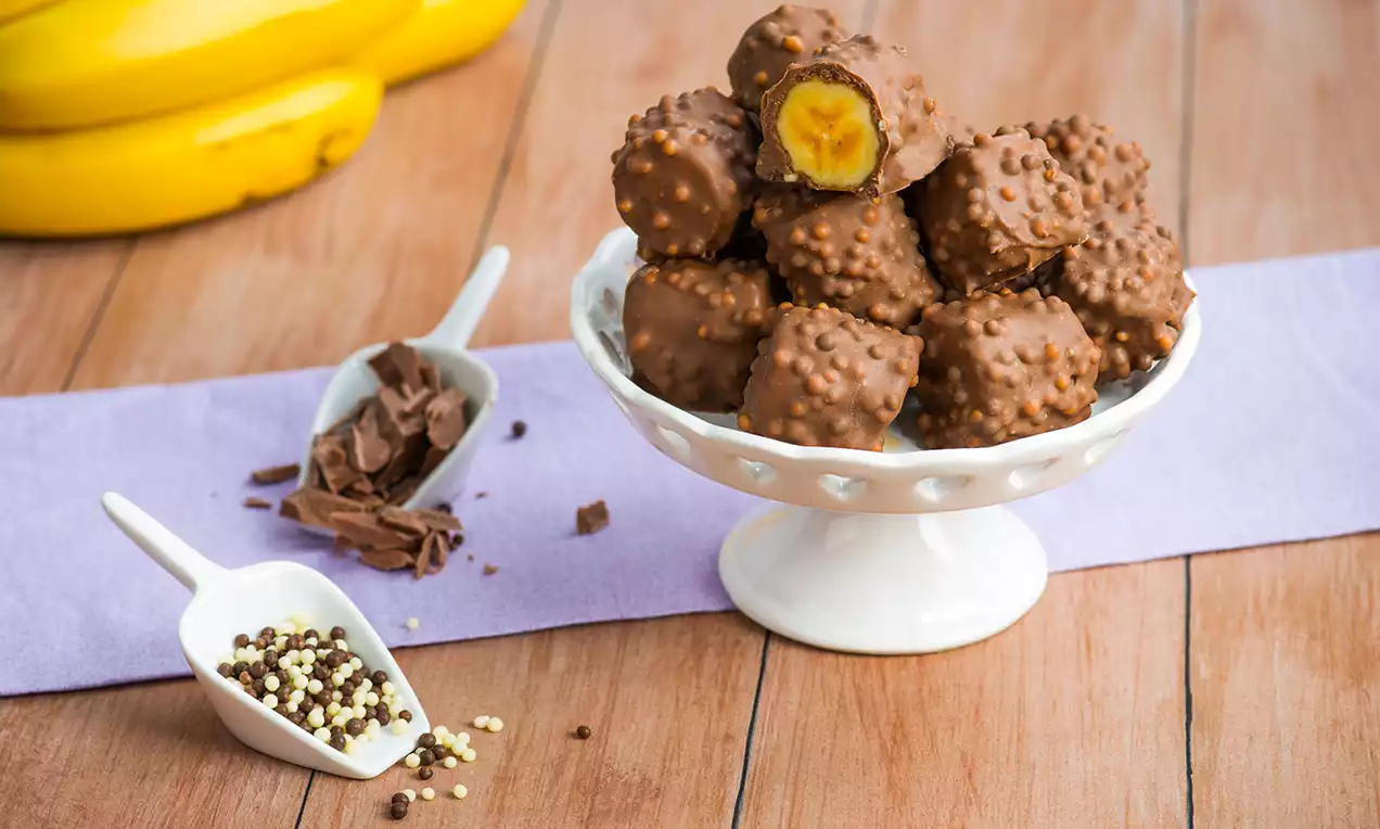 Biter av sprø krokantrukket banan i brun sjokolade med nøttebiter, lagt på et hvitt fat, omgitt av lilla tekstil og dekor sjokoladekuler